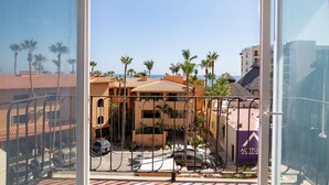 A view from a balcony overlooking a street with parked cars, palm trees, and buildings with red-tiled roofs.