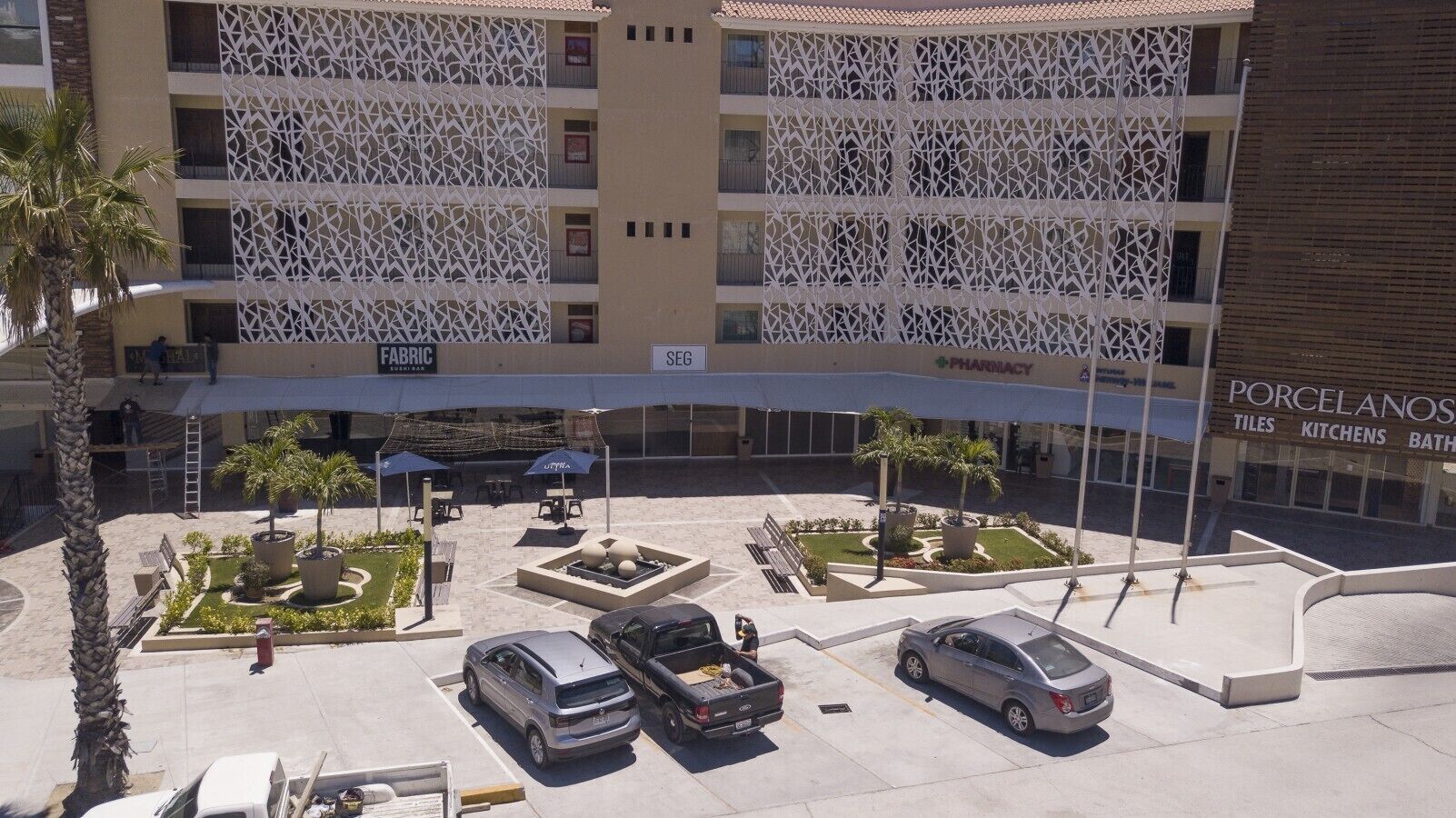 Een parkeerplaats met meerdere auto's, een groen middeneiland met palmbomen en een gebouw met een gebeeldhouwd gevel, borden met 'FABRIC' en 'PORCELANOS TILES KITCHENS BATHROOMS'.
