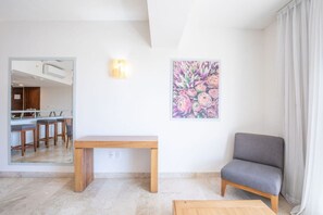 A living room with a grey sofa, a wooden coffee table, a painting of flowers on the wall, and a kitchen visible through an open door.