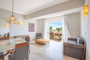 Ein modernes Wohnzimmer mit einem Glastisch, grauen Sofas, einem Holztisch und einem Balkon mit Blick auf Palmen.