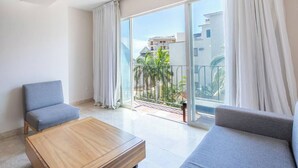 Una sala de estar moderna con una mesa de centro de madera, dos sofás grises y un gran ventanal corredero que da a un balcón con vistas a edificios y palmeras.