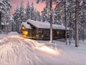 Una cabana de fusta amb un teulat cobert de neu i un interior encesa, envoltada d'arbres carregats de neu i un camí ple de neu.