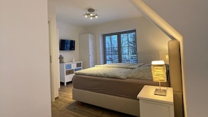 Een moderne slaapkamer met een groot bed, een nachtkastje met een lamp, een televisie en een raam met uitzicht op bomen.