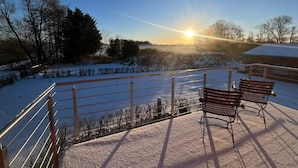 Een besneeuwde buitenruimte met banken en een hekwerk met uitzicht op een landschap met bomen en een gebouw op de achtergrond.