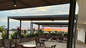 An outdoor seating area with tables and chairs, a round wooden table, and a view of the sunset over a cityscape. - Modern 2BR Retreat | Pool, Wi-Fi, AC & Prime Location, 10 Min from Everything (Puerto Vallarta)