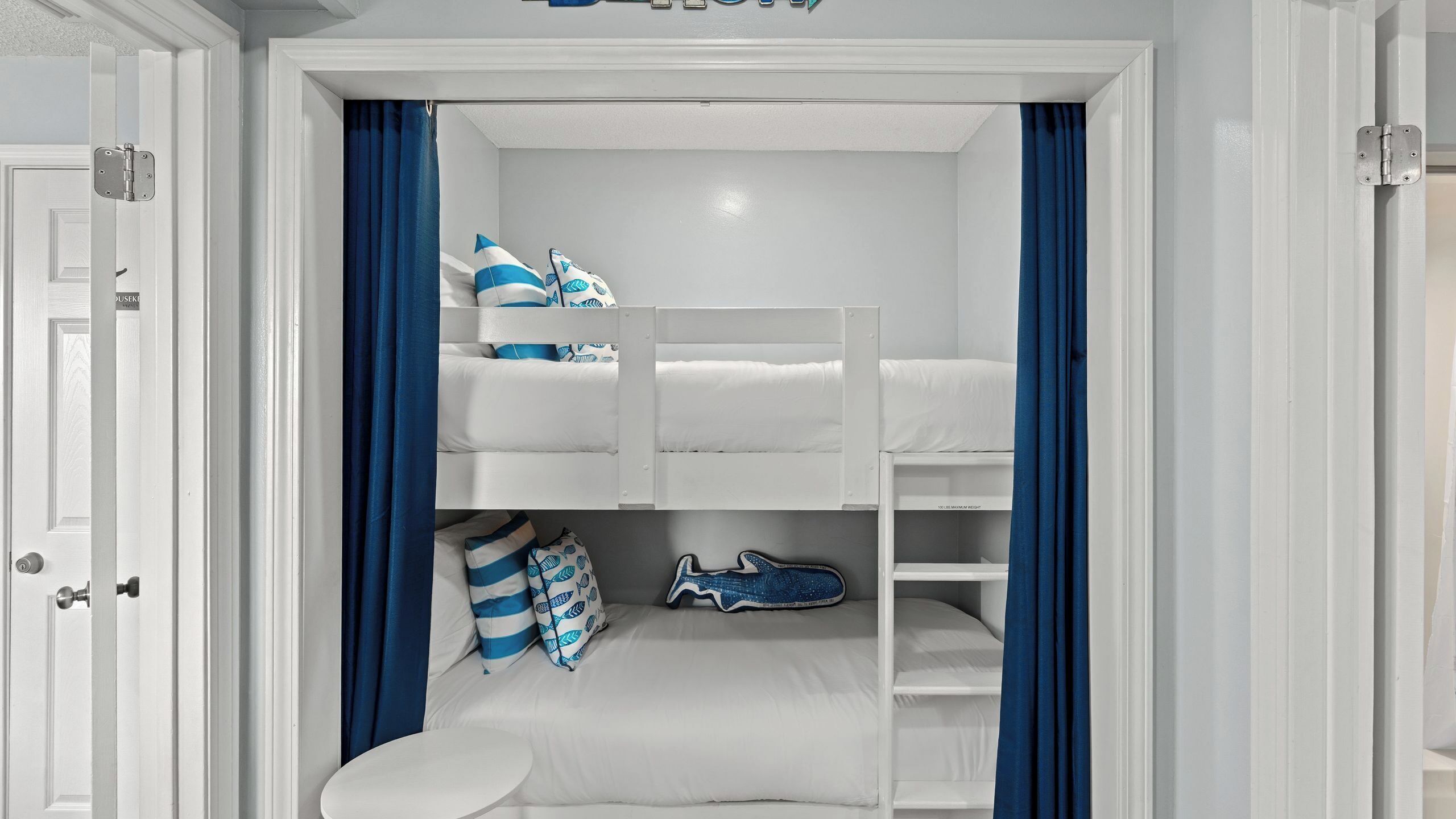 Een slaapkamer met stapelbed, een witte deur, blauwe gordijnen en een klein wit tafeltje.