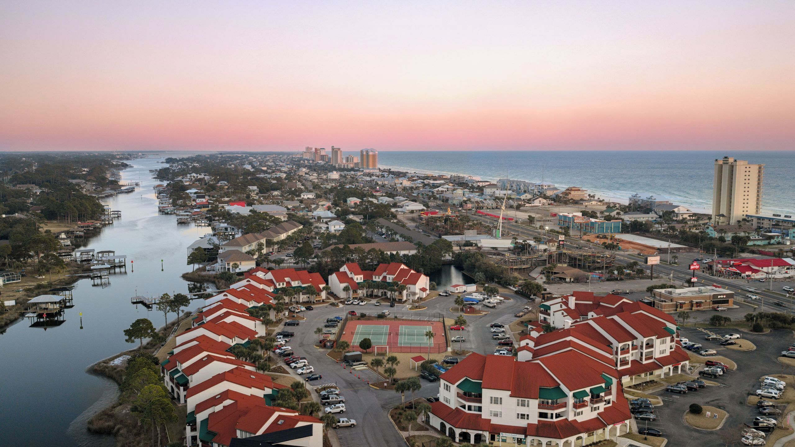 An aerial view of a coastal town with a marina, residential buildings, and a prominent hotel with a red roof.