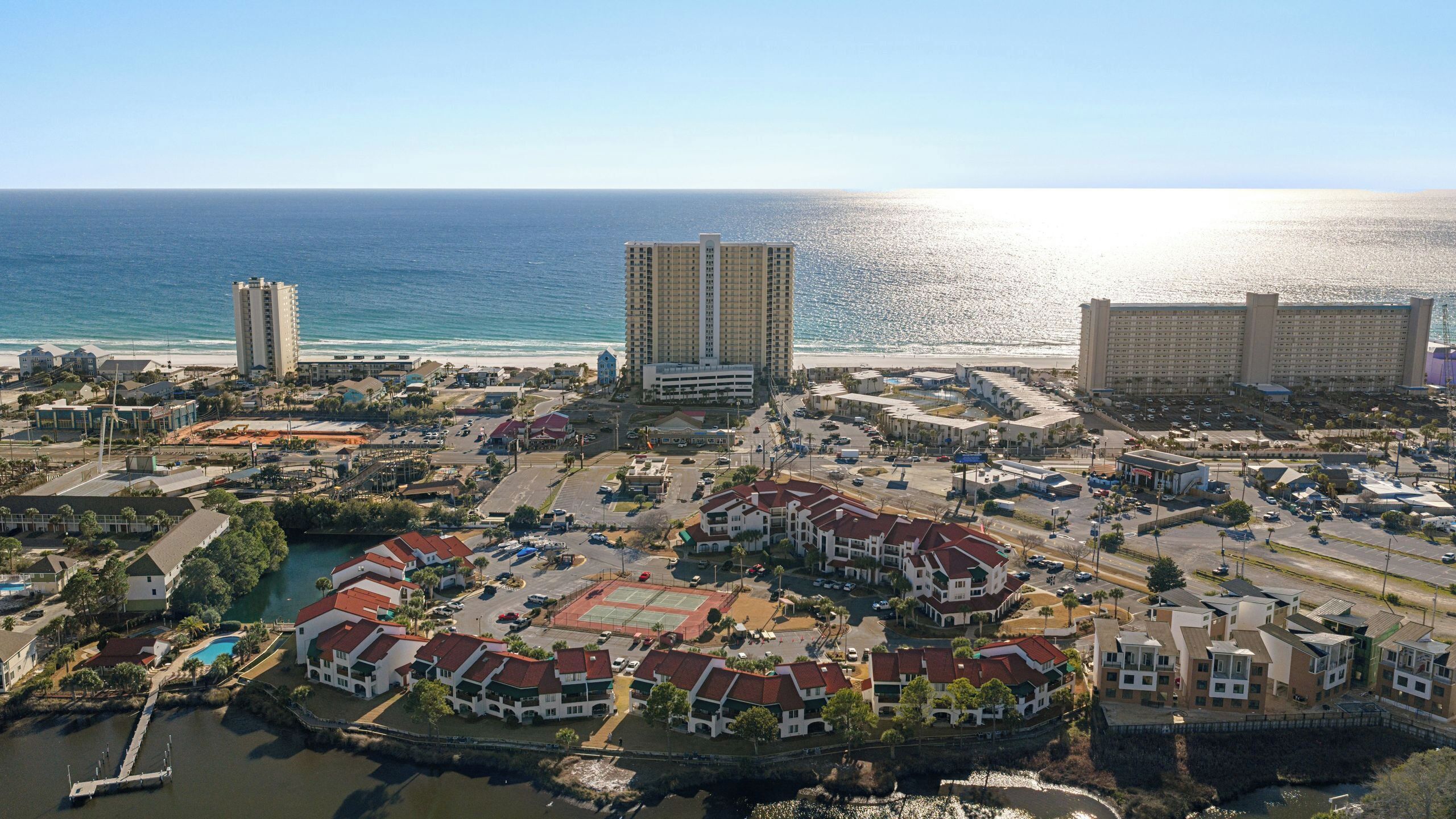 Vista aérea de un complejo turístico frente al mar con hoteles en la playa, un puerto deportivo y un centro deportivo.