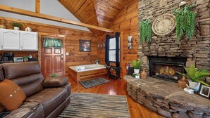 A cozy cabin interior with a stone fireplace, wooden walls, a leather sofa, and a small bathroom. - Hot Tub! Location! Cozy Studio Log Cabin,Fireplace (Gatlinburg)