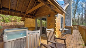 A wooden deck with a hot tub, two rocking chairs, and a stone wall. - Hot Tub! Location! Cozy Studio Log Cabin,Fireplace (Gatlinburg)