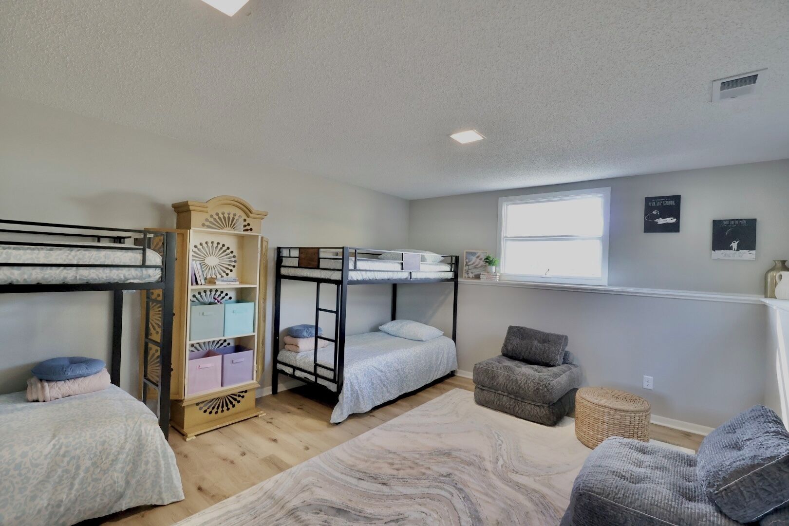 A dormitory room with bunk beds, a wooden cabinet, a small armchair, and a window with artwork.