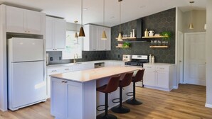 Cocina moderna con armarios blancos, una isla central con taburetes de bar, luces colgantes y un salpicadero con baldosas hexagonales.