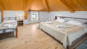 Ein gemütliches Schlafzimmer im Dachgeschoss mit zwei Betten, Holzbalkendecke, einem kleinen Fenster mit Kreuz und einem Nachttisch aus Holz mit Lampe.
