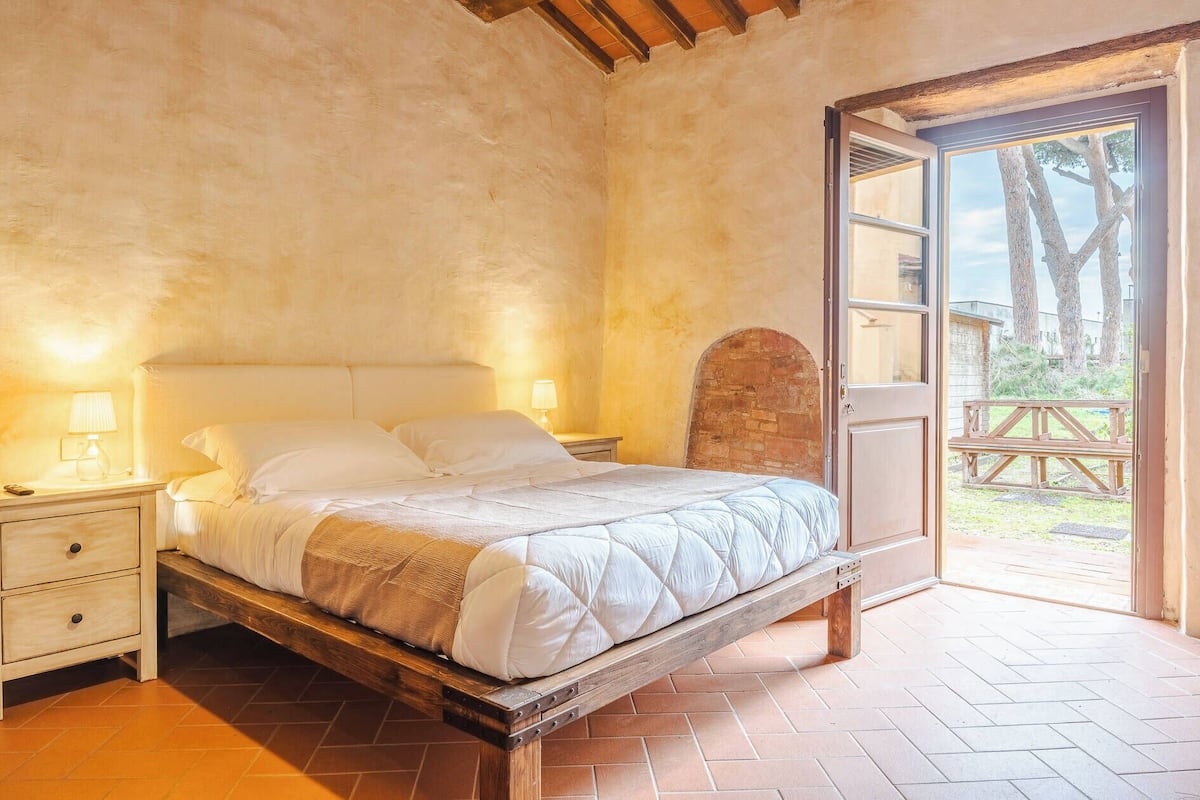 A bedroom with a wooden bed frame, a nightstand with a lamp, and a view of an outdoor area with a wooden deck and trees.