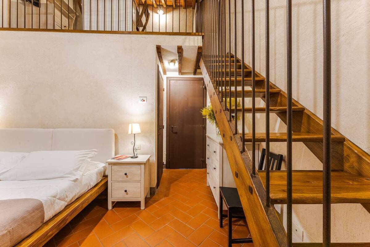 A bedroom with a wooden bed, a nightstand with a lamp, a dresser, and a staircase with wooden railings.