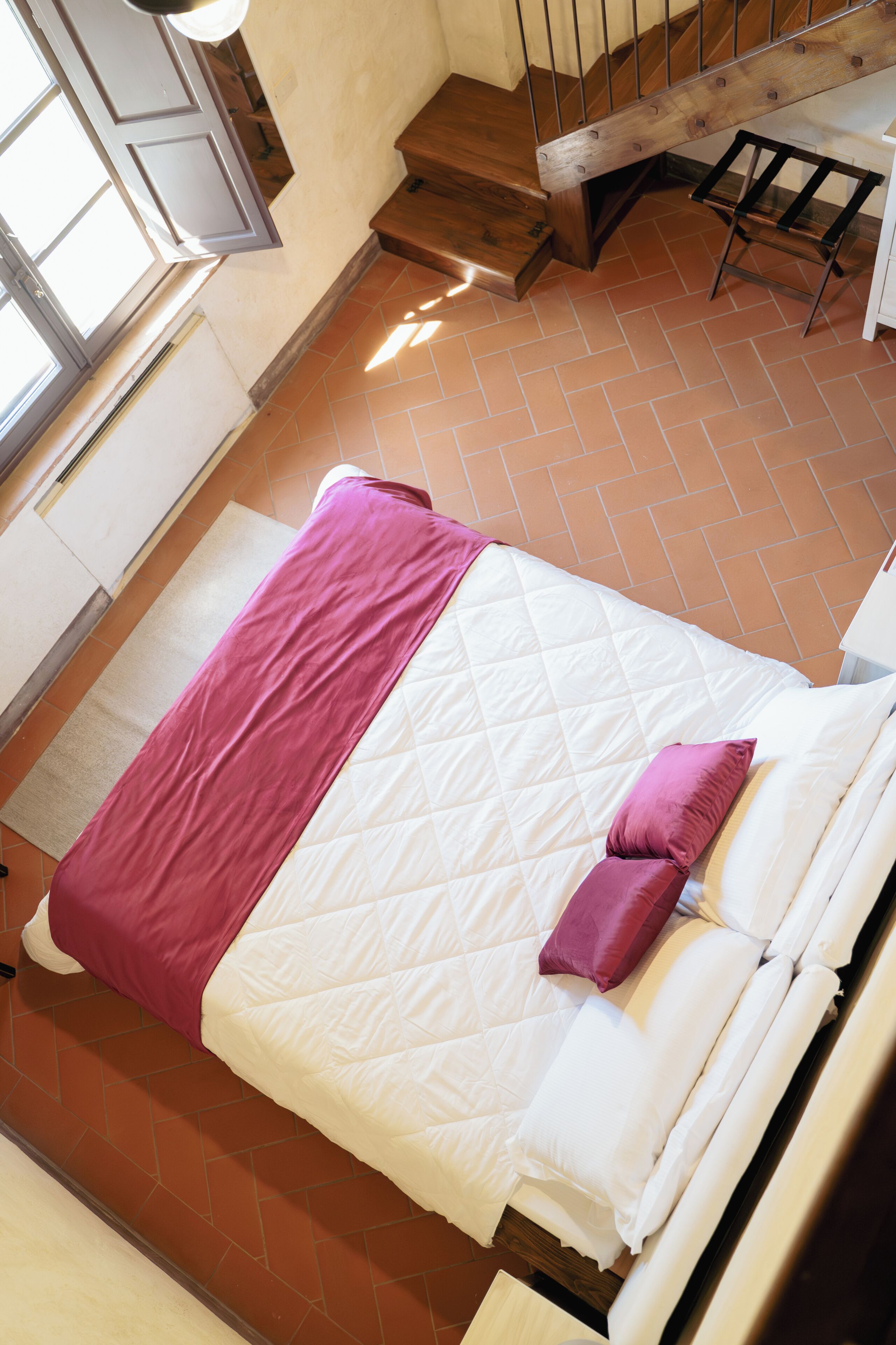 Een bed met een gewatteerd wit matras, een roze deken en een paars kussen in een kamer met een betegelde vloer en een houten trap.