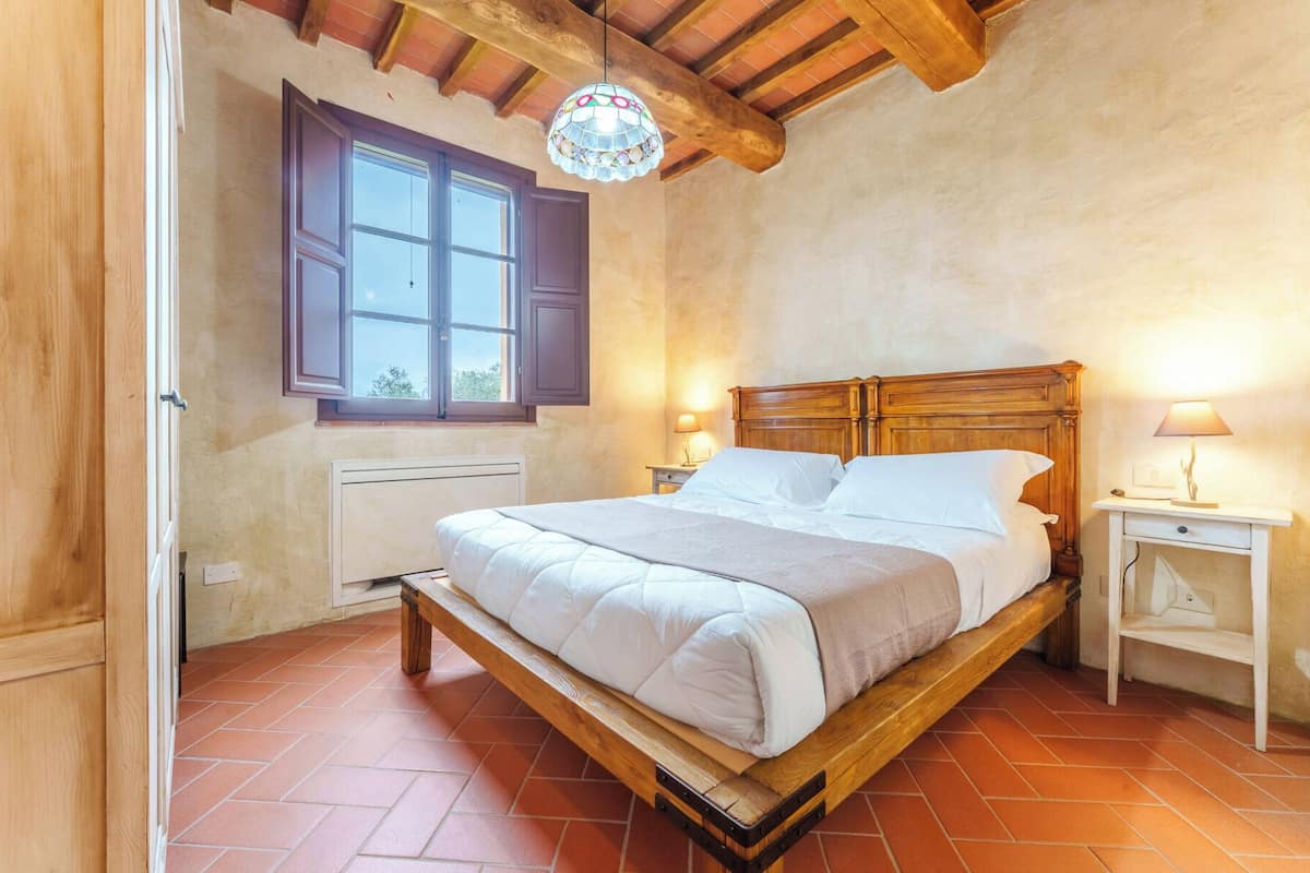 A bedroom with a wooden bed, a window with shutters, a radiator, and a nightstand with a lamp.