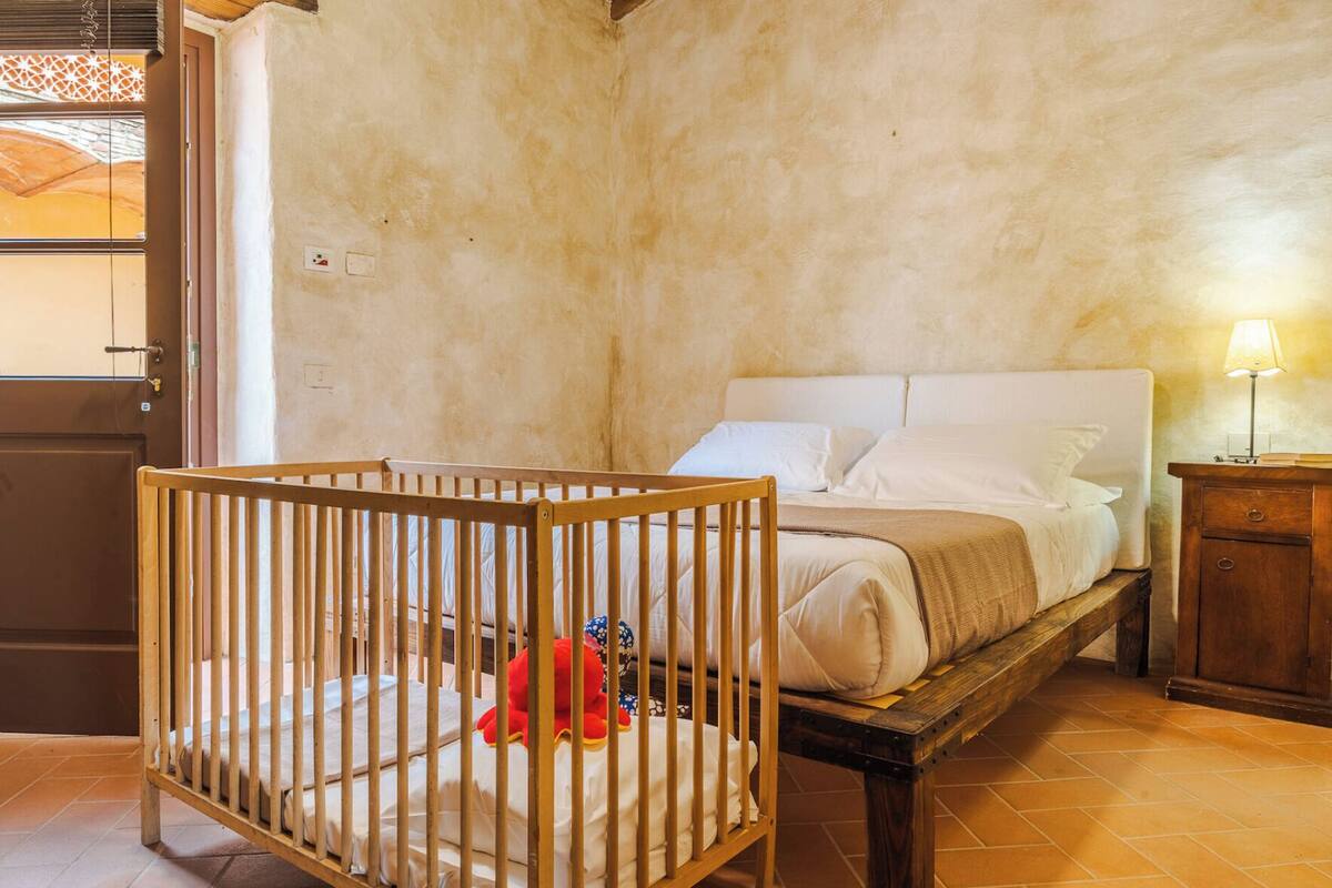 A room with a crib, a bed with white linens, a wooden nightstand, and a lamp.