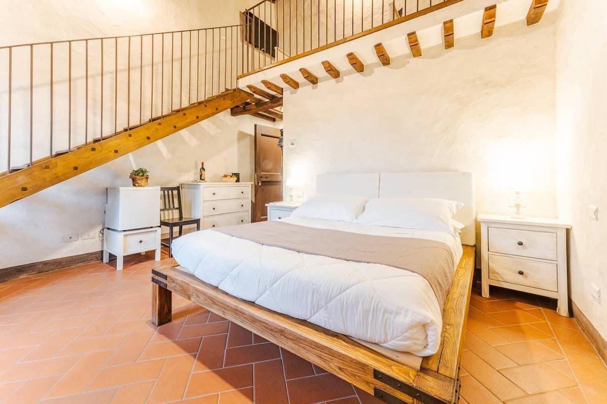 A bedroom with a wooden bed, a white nightstand, a wooden chair, a small table, and a staircase.