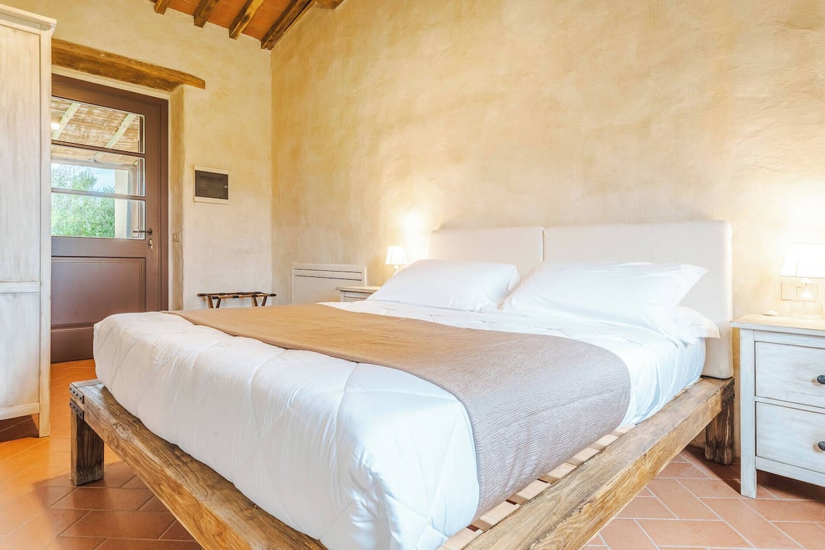 A bedroom with a wooden bed frame, white bedding, a nightstand with a lamp, and a door leading outside.