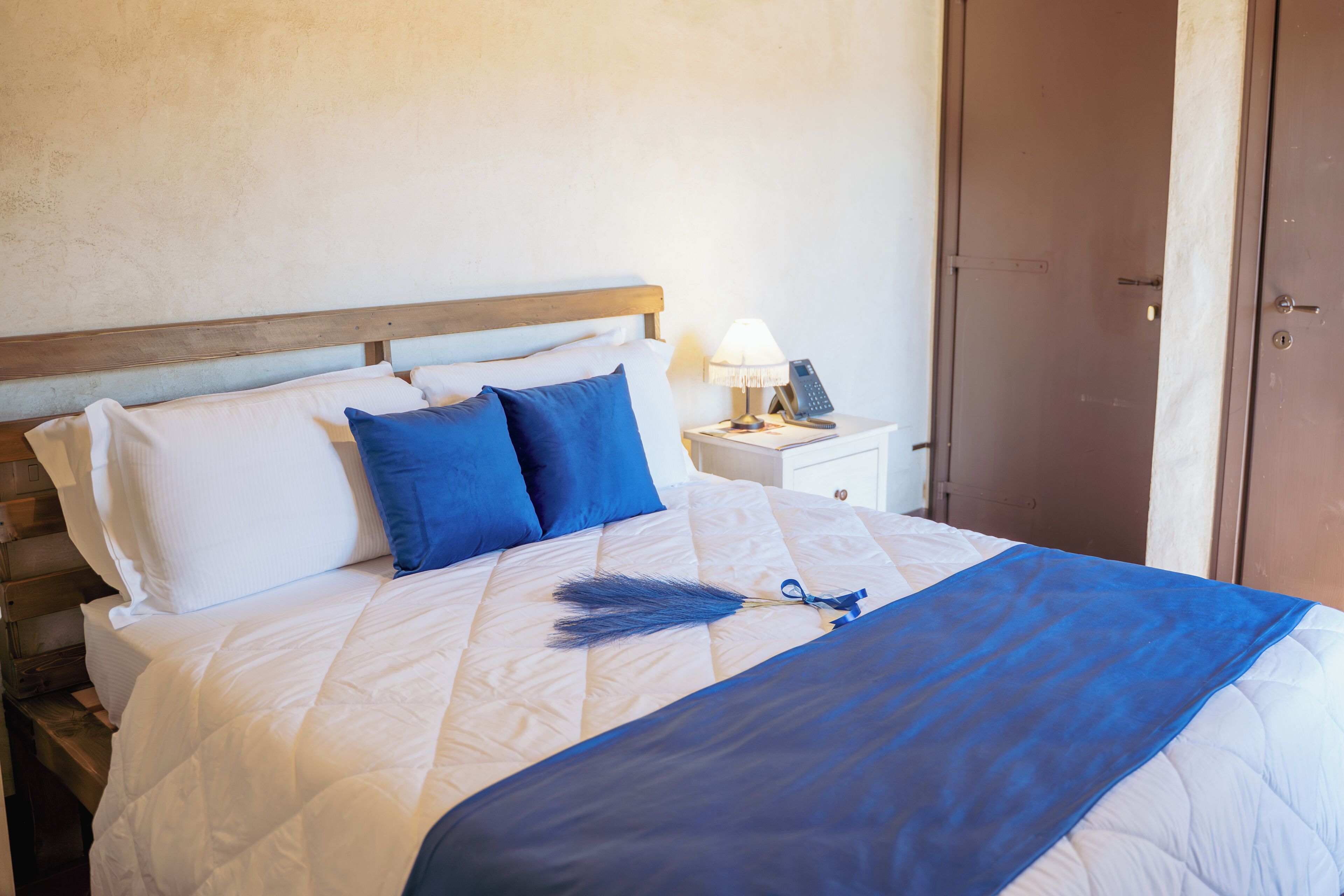 Un lit avec une tête de lit en bois, des oreillers blancs et bleus, un plaid bleu, une lampe de chevet et un téléphone sur une table de chevet.