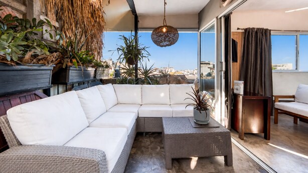 Un balcone moderno con un divano angolare bianco, un tavolino da caffè in vimini e piante in vaso.