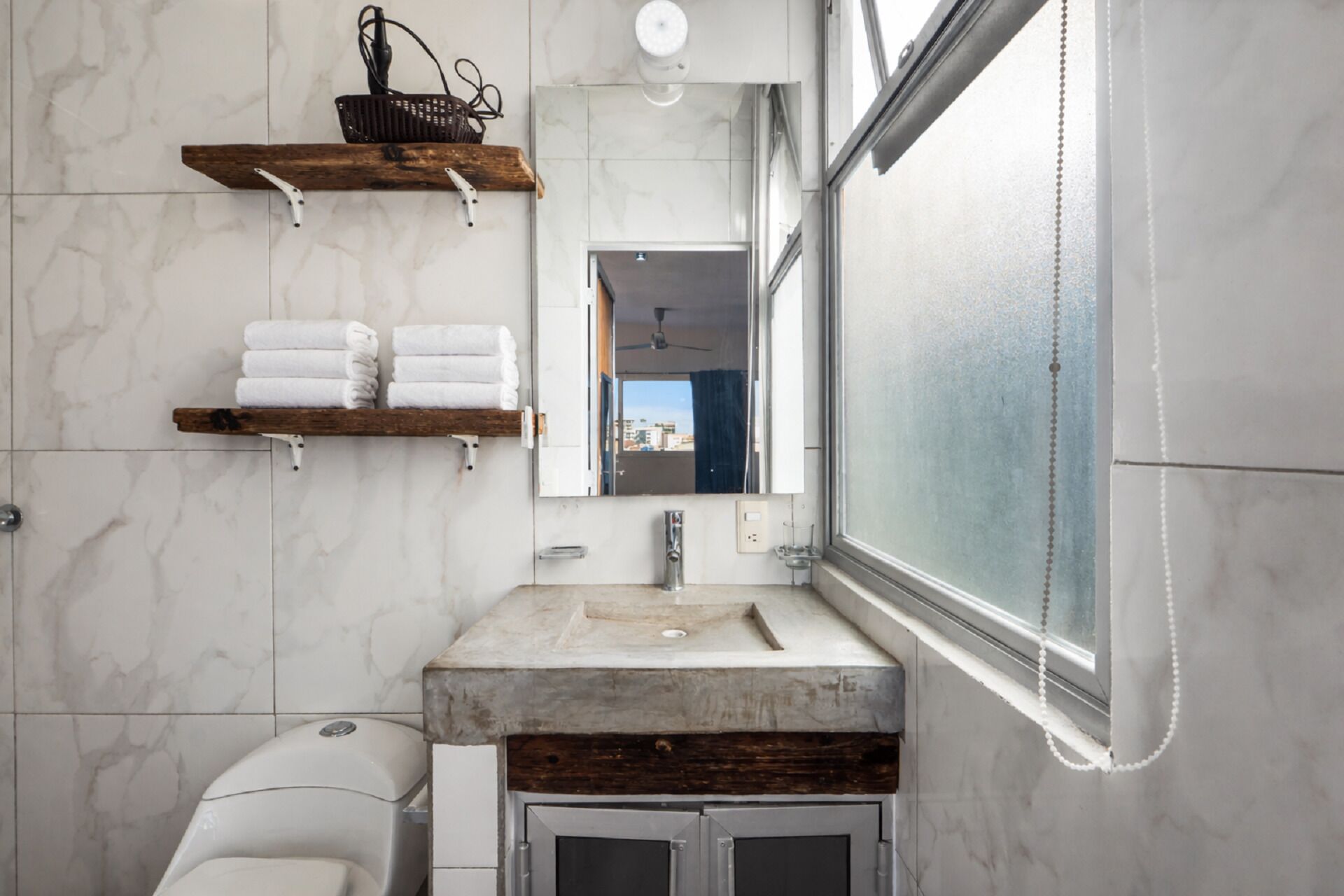 Un baño con pared de mármol, un estante de madera con toallas, un espejo y una ventana.