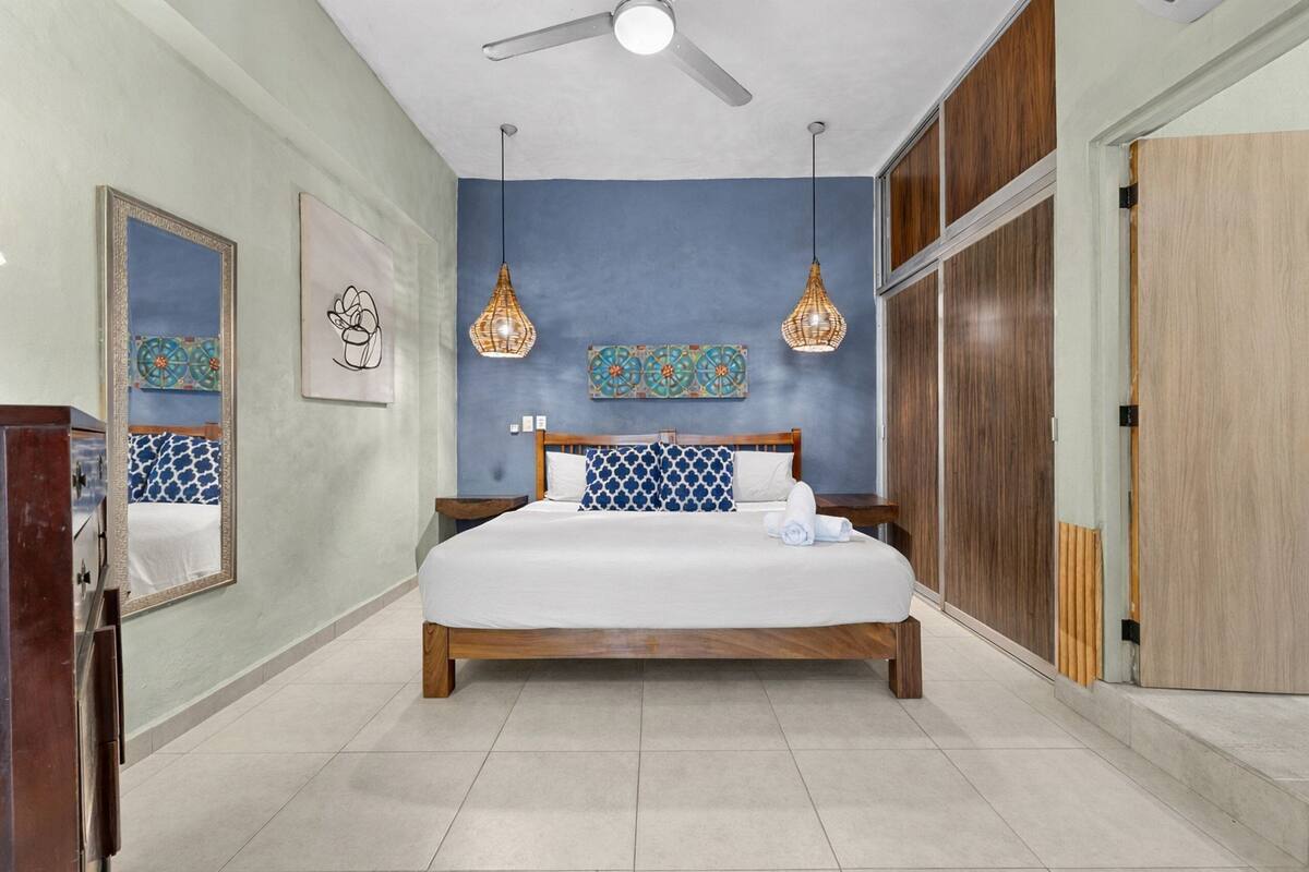 A bedroom with a bed, ceiling fan, pendant lights, artwork, and a wardrobe.