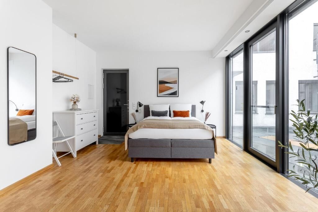 Une chambre moderne avec un grand lit, une commode blanche, un miroir et une grande fenêtre donnant sur l’extérieur.