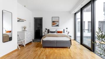 Une chambre moderne avec un grand lit, une commode blanche, un miroir et une grande fenêtre donnant sur l’extérieur.