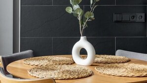 A round wooden table with woven placemats, surrounded by grey upholstered chairs. A white vase with green foliage is placed on the table. - Global Living Augsburg Rathausplatz (Augsburg)
