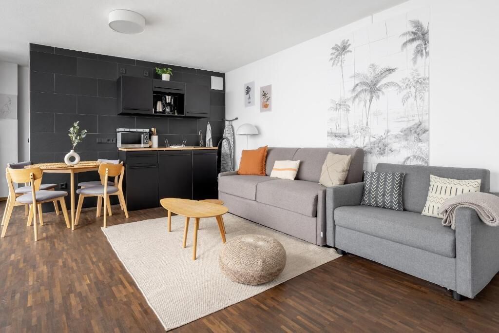A modern living room with a grey sofa, wooden chairs, a small wooden table, and a kitchen area with black cabinets and a microwave.