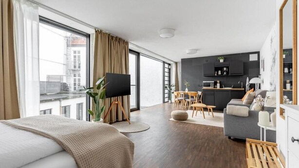 A modern hotel room with a large bed, a dining area, a kitchen, and a view of the city. - Global Living Augsburg Rathausplatz (Augsburg)