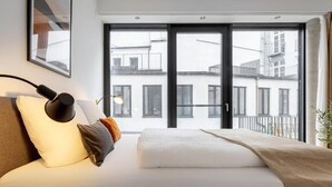 A modern bedroom with a large window, a bed with white bedding, a black lamp, and a view of buildings outside. - Global Living Augsburg Rathausplatz (Augsburg)