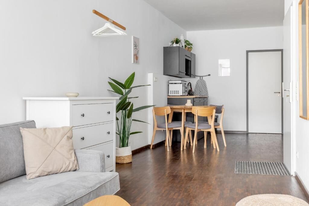 Una sala de estar moderna con un sofá gris, una mesa de comedor con sillas, una zona de cocina con microondas y una planta en maceta.