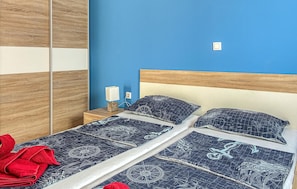 Una cama doble con cubrecama con motivos náuticos, un cabecero de madera, una mesita de noche con lámpara y una toalla roja en el suelo.
