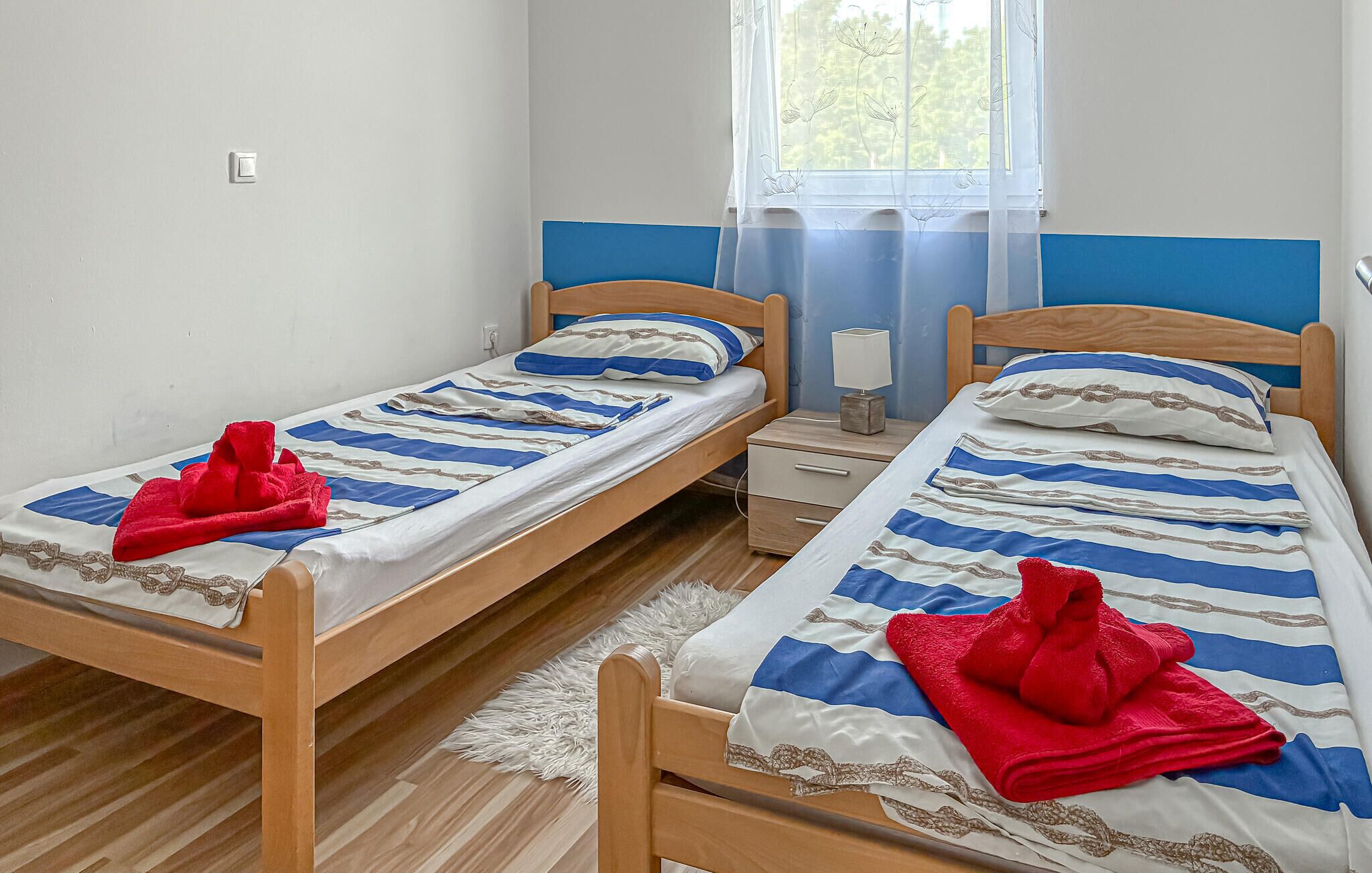 A room with two single beds, a nightstand with a lamp, a window with a sheer curtain, and a white rug.