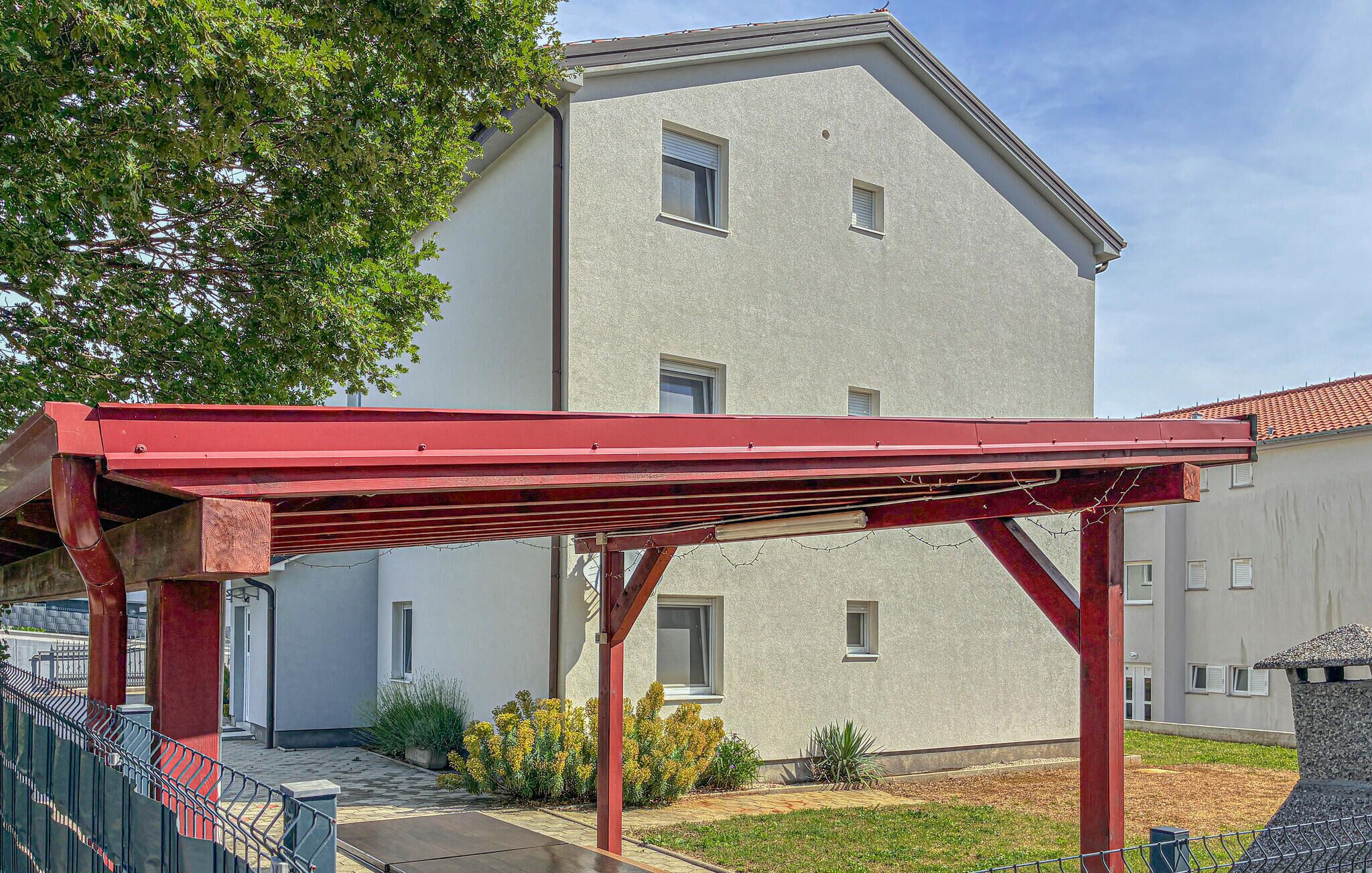 Rakennuksessa on punainen metallirunko, mahdollisesti pergola, ja se sijaitsee kaksikerroksisen talon edessä. Talossa on harmaa julkisivu ja useita ikkunoita.