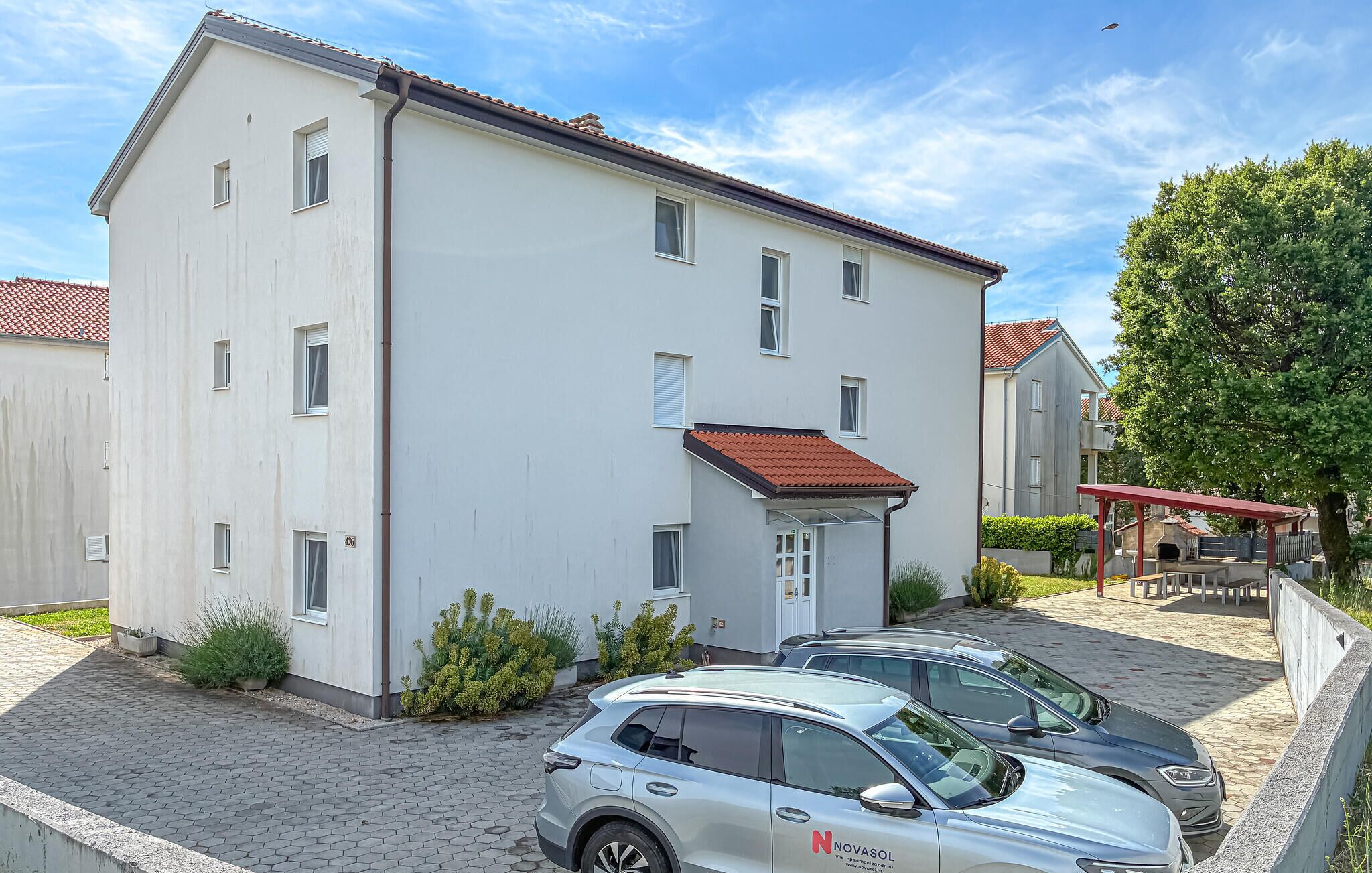 Un edificio a due piani con tetto in tegole rosse, un ingresso coperto e un'area di parcheggio con tre auto.