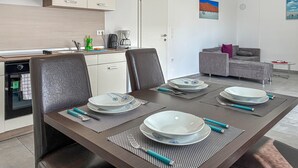 Een eetgedeelte met een tafel voor vier personen, een keuken met moderne apparatuur, een grijze bank en kunstwerken aan de muur.