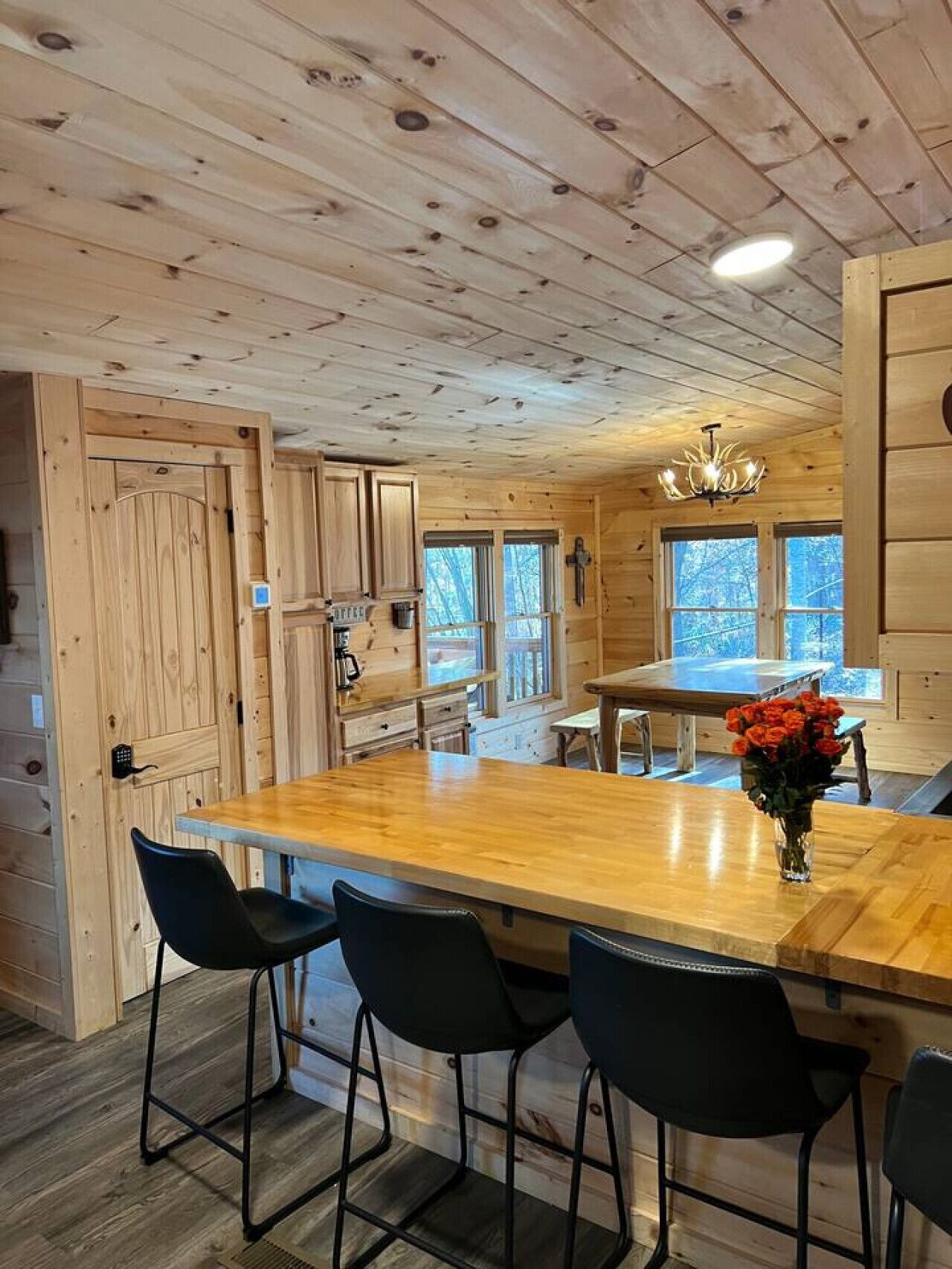 L’intérieur d’un chalet en bois comprend une grande table à manger, quatre chaises noires, un lustre et une fenêtre donnant sur les arbres.