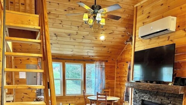 Ein Holzhütten-Innenraum mit Kamin, einem darüber montierten Fernseher, einem Essbereich mit Stühlen und einer Treppe mit einer Lampe auf der Zwischenebene.