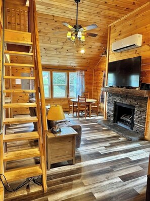 A wooden cabin interior with a fireplace, a TV mounted above it, a dining area with chairs, and a staircase with a lamp on the landing. - Creekside Blessings 
Cabin (Beech Mountain)