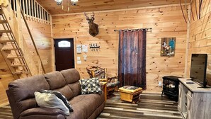 Interior de uma cabana de madeira com um sofá marrom, um baú de madeira, uma lareira e uma cabeça de veado na parede.