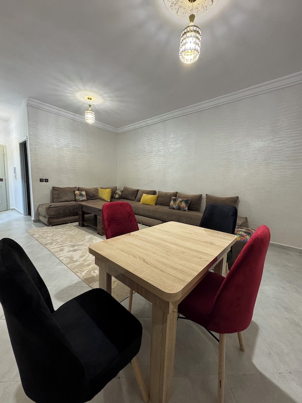 A modern living room with a wooden dining table, red and black chairs, a grey sectional sofa with colorful cushions, and two crystal chandeliers.