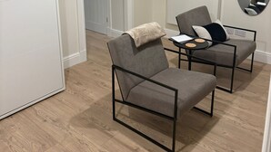 Une chambre moderne avec un plancher en bois, une armoire blanche, un fauteuil gris avec un plaid, une petite table avec une lampe et un miroir au mur.