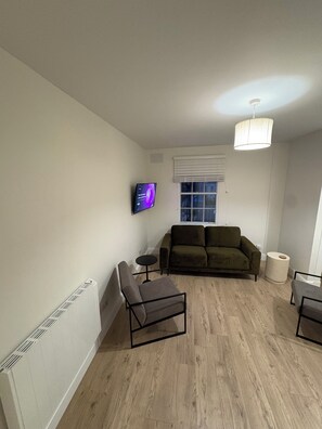 Uma sala de estar com sofá, uma mesa pequena, uma cadeira, TV fixada na parede, janela com persianas e luminária no teto.