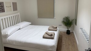 Un lit bien fait, avec du linge de lit blanc et deux serviettes pliées, une plante en pot, un miroir et une grille d’aération au mur.