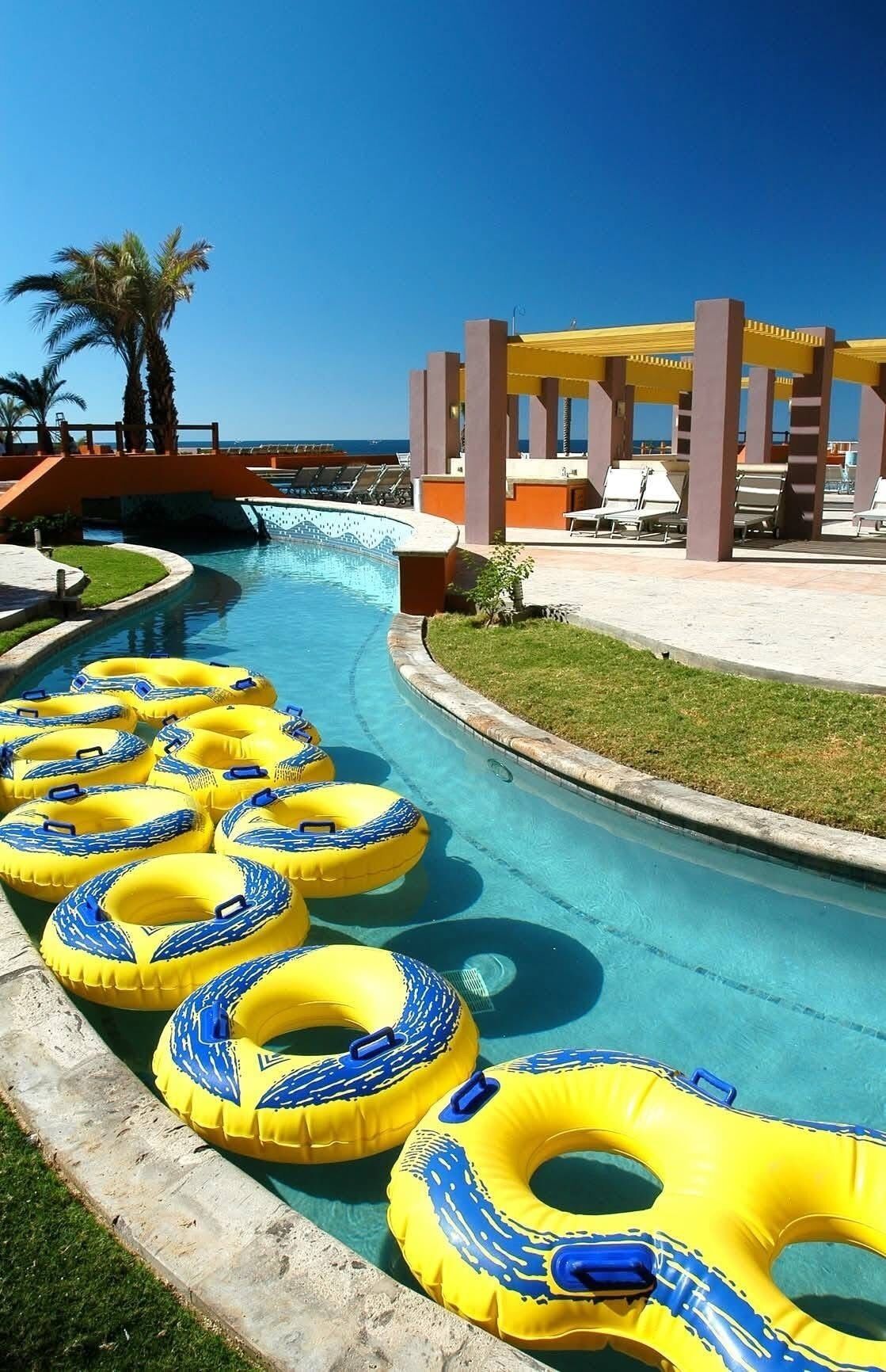 A pool area with yellow and blue inflatable rings, a palm tree, and a covered seating area with a view of the ocean.