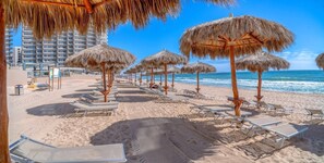 A beach with rows of sun loungers and thatched umbrellas, a clear blue sky, and a building in the background. - 3BR Ocean View Condo, Las Palomas Resort, Pools, Lazy River & Water Slide (Puerto Peñasco)